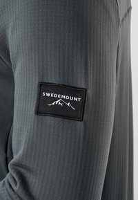 Manche en tissu gris texturé avec un patch rectangulaire noir portant l'inscription "SWEDEMOUNT" au-dessus d'un logo de montagne blanc.