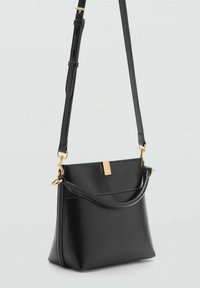 Čierna kožená crossbody taška s nastaviteľným popruhom, zlatými hardvérovými detailmi a tenkým predným uchom, umiestnená na jednoduchom svetlom pozadí.