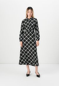 Une femme se tient contre un fond blanc, vêtue d'une robe midi à manches longues à motifs diamants noir et blanc et de chaussures à talons ouverts noires à bout pointu.
