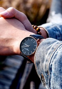 Reloj con esfera negra, marcadores de horas delgados en blanco, caja dorada y correa de cuero negra. Lleva puesto en una muñeca con una pulsera de cuentas de madera.