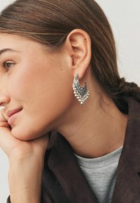 Sølvøreringe med halvmåneform, indviklet sort detaljering og hængende perleaccenter. Båret på en persons øre.