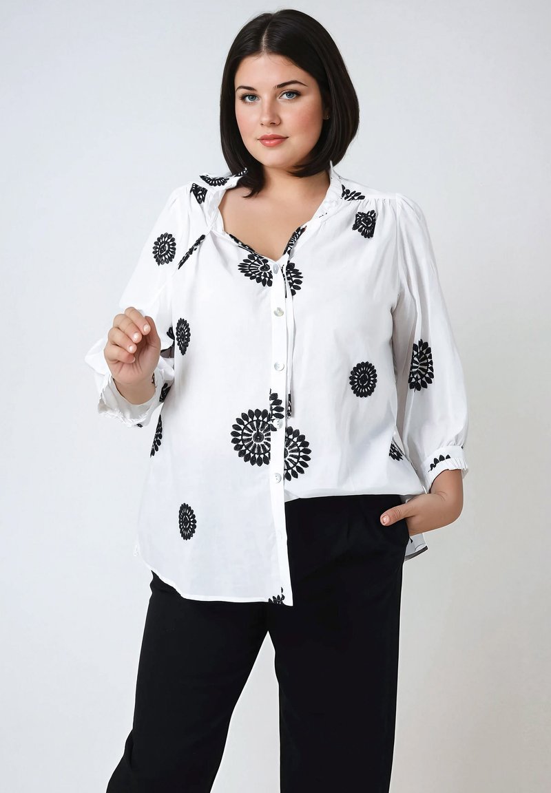 Witte shirt met zwarte bloemige borduursels, knopen aan de voorkant en lange mouwen. Gekleed met zwarte broeken. Glad stof, ontspannen pasvorm en subtiele details.
