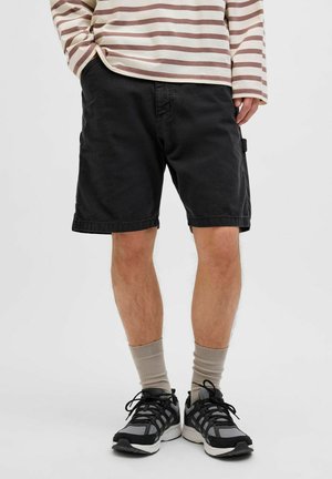 Zwarte shorts met een ontspannen pasvorm, voorzien van zijzakken en een zoom die boven de knie eindigt. Draagt beige sokken en zwarte sportschoenen.