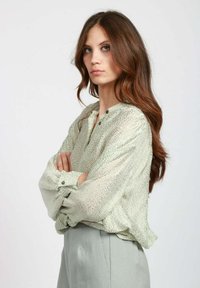 Blusa verde chiaro, con texture e vestibilità rilassata, maniche lunghe, dettagli a bottoni e un motivo sottile, abbinata a pantaloni grigi a vita alta.