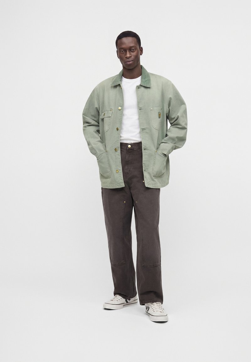 Mann trägt eine hellgrüne, geknöpfte Jacke, weißes T-Shirt, dunkelbraune Hose und weiße Turnschuhe, steht vor einem schlichten hellen Hintergrund.