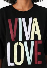 Juodas trumpas rankovės marškinėlis, kuriame ryškiomis, daugiamačių spalvų raidėmis užrašyta "VIVA LOVE" skirtingais aukščiais ir stiliais, su tekstūruotu paviršiumi.