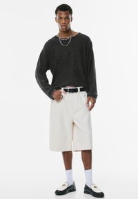 Zwarte ribbed shirt met lange mouwen, witte gestreepte shorts, zwarte riem, witte sokken, zwarte instappers met accent. Neutrale achtergrond.