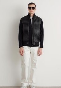 Μαύρο μπουφάν με επενδεδυμένο μπροστινό μέρος, φερμουάρ και ψηλό γιακά. Συνδυάζεται με λευκά παντελόνια και αθλητικά παπούτσια. Το μοντέλο φοράει σκούρα γυαλιά ηλίου.