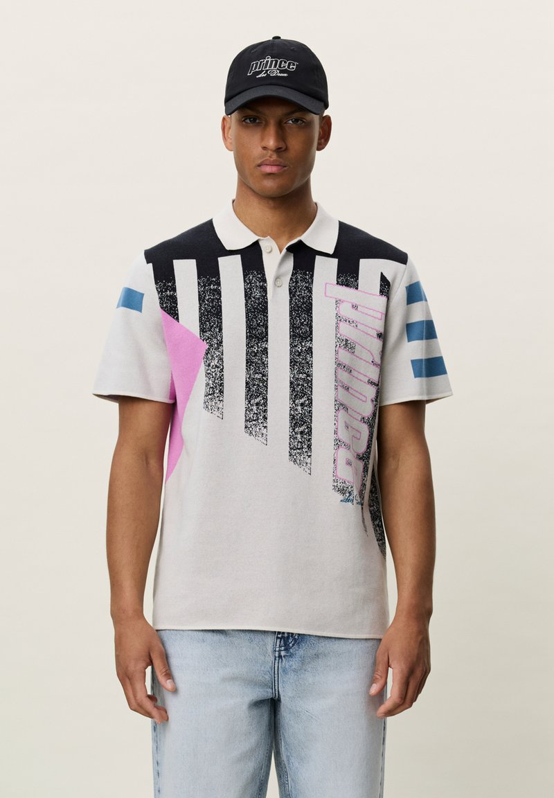 Jeune homme portant une casquette noire et un polo blanc avec des motifs géométriques noirs, roses et bleus et un jean bleu clair.