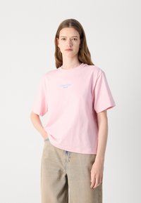 T-shirt di cotone rosa con maniche corte, scollatura rotonda e logo blu di Calvin Klein Jeans sulla parte anteriore. Indossato con pantaloni in denim chiaro.