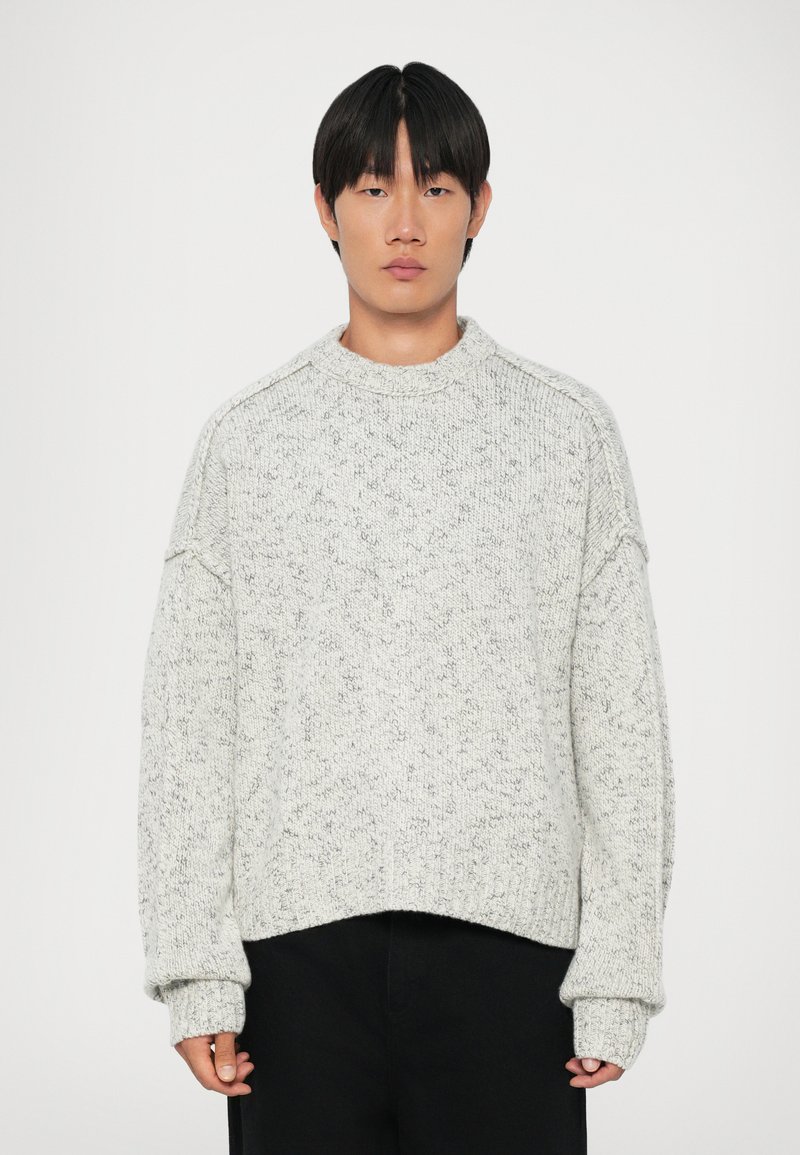 Lys grå chunky strikkegenser med høy hals, senkede skuldre og ribbestrikkede mansjetter. Teksturen har et subtilt prikket mønster.