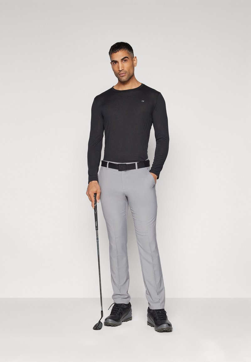Svart langermet topp, lys grå bukser med belte, svarte sko, og en golfklubb. Uniform design og tettsittende silhuett.