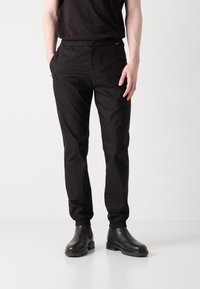 Sorte elastiske bukser med sidelommer, tapered fit og strækbare afslutninger. Bæres med sorte ankelstøvler på en neutral baggrund.