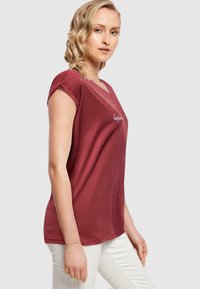 Rød t-skjorte med kort erme og v-hals, med teksten "Inspire." i hvitt. Stoffet ser mykt og lett ut. Matchet med hvite bukser.