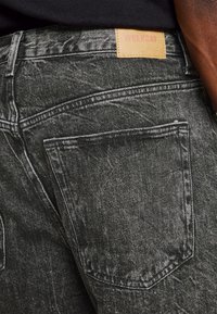 Vaqueros de mezclilla negra con una textura desgastada, que presentan un parche de cuero marrón claro en la cinturilla etiquetado como "WEEKDAY" y bolsillos traseros estándar.