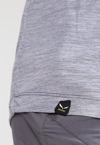 Šedé, texturované tričko z látky s malou černou etiketou v bílých akcenty na lemu, zobrazující logo značky.