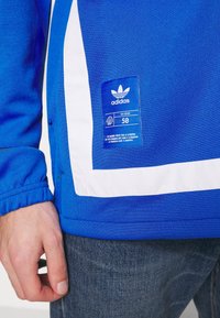 Sininen collegepaita valkoisilla yksityiskohdilla, resorihihansuuilla ja etiketillä, jossa Adidas-logo ja koko 50. Kankaan pinta näyttää sileältä.