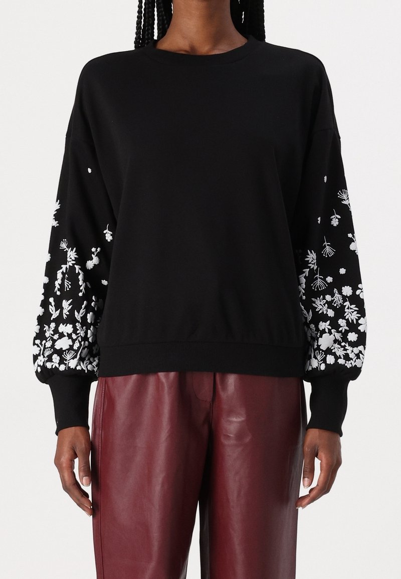 Personne portant un pull noir avec des motifs floraux blancs sur les manches et un pantalon en cuir bordeaux sur un fond uni.