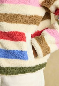 Osoba nosi viacfarebnú pruhovanú fleecovú bundu s rukou v bočnom vrecku, zobrazujúcu textúru a útulný dizajn.