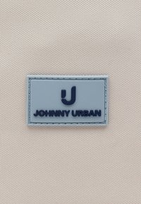 Etiqueta rectangular gris con un "J" estilizado encima del texto "JOHNNY URBAN" sobre un fondo de tela beige texturizada.