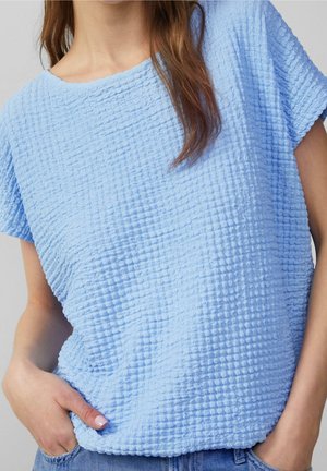 Nahaufnahme einer Person, die ein strukturiertes hellblaues kurzärmeliges Oberteil trägt, mit den Händen in den Taschen und sichtbarem lockeren braunen Haar.