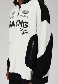 Melnas un baltas vilnas pulovers ar apģērbtu sānu kabatu, lielu "RACING" tekstu un klātienē pārbaudītu rakstu akcents priekšpusē.