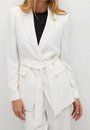 Kobieta ubrana w biały żakiet z paskiem na czarnym topie, stojąca na jednolitym białym tle.