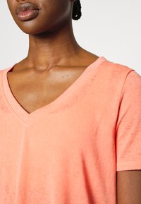 T-shirt cor-de-rosa claro com decote em V, apresentando mangas curtas, textura suave e um ajuste descontraído, destacando um acabamento de tecido liso e uniforme.