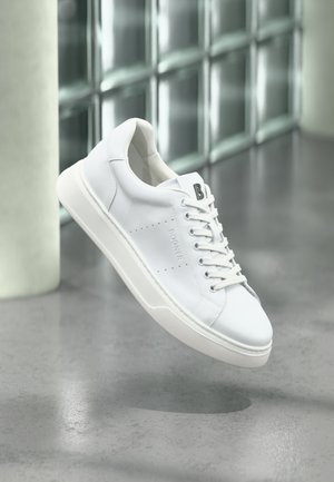 Sneaker bianca bassa con lacci, pannello laterale traforato e logo "B" sulla linguetta, sospesa sopra un pavimento grigio lucido vicino a blocchi di vetro.