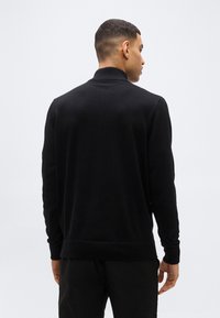 Άνδρας με κοντά μαλλιά, που φοράει μαύρο πουλόβερ με λαιμόκοψη και μαύρο παντελόνι, γυρισμένος με την πλάτη προς μια απλή φωτεινή επιφάνεια.