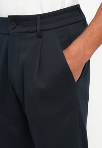 Pantaloni a pieghe blu navy con una texture liscia, tasche laterali e chiusura con bottone in vita. Retro elastico per il comfort.