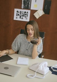 Cardigan gris flou avec des boutons, tenant une tasse transparente contenant un liquide sombre. Téléphone ancien blanc et ordinateur portable sur un bureau avec des carnets.