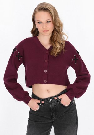 Ung kvinde med bølget blondt hår iført en cropped bordeaux cardigan med knapper og sorte blonder detaljer samt sorte højtaljede jeans, hænderne i lommerne.