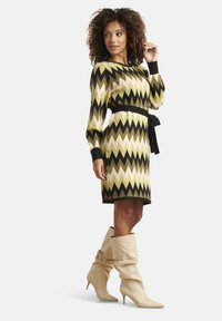 Vestito in maglia con motivo a zigzag verde, beige e nero, maniche lunghe e vita annodata. Abbinato a stivali morbidi color crema.