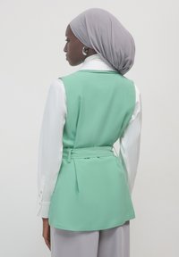 Mintgrüner Weste mit definierter Taille, übereinander getragen über einer weißen Bluse. Verfügt über eine glatte Textur und ein zurückgebundenes Design. Grauer Hijab betont das Gesamtbild.