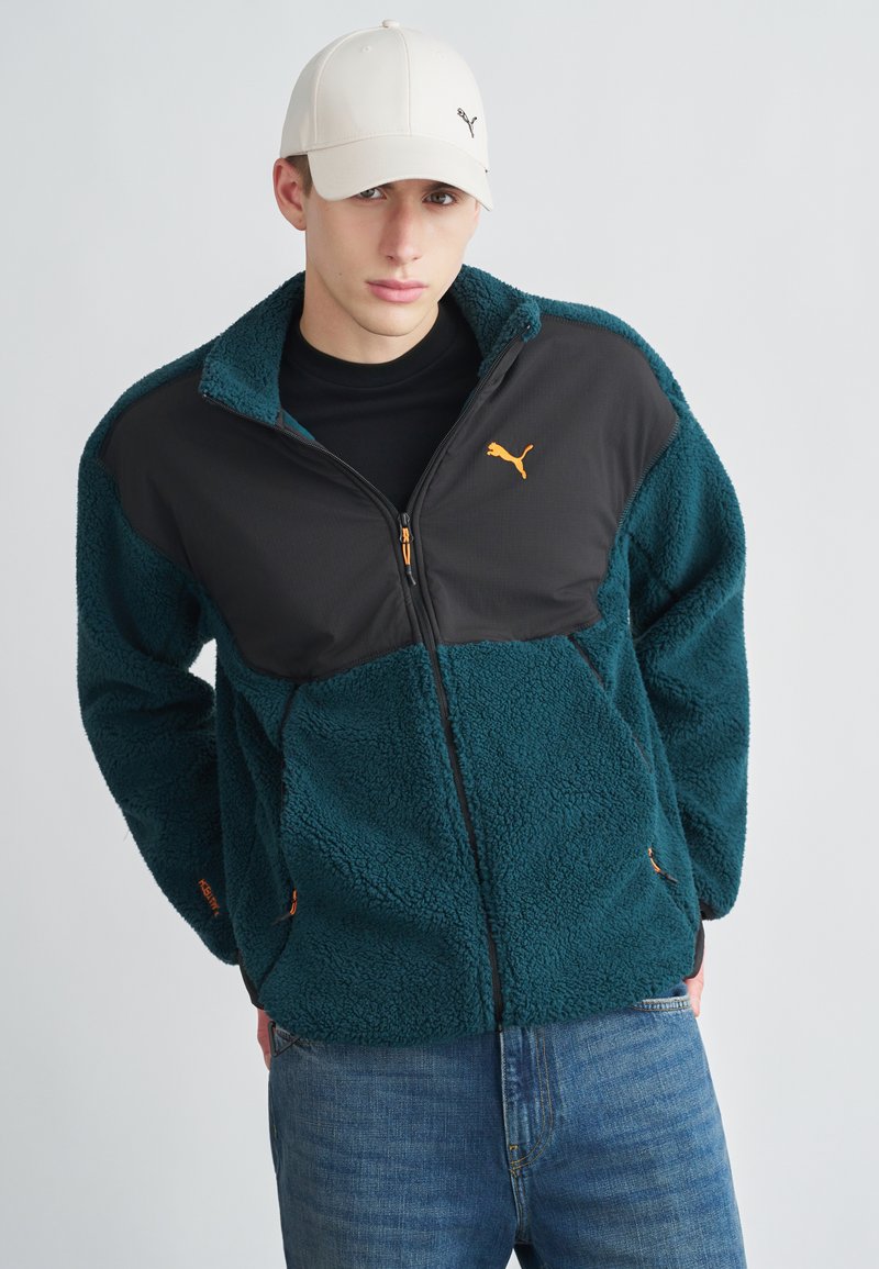 Vihreä fleecetakki mustilla paneeleilla, oransseilla yksityiskohdilla ja etuvetoketjulla. Yhdistetty valkoiseen lippalakkiin, jossa on logo. Vaaleansiniset farkut päällä.