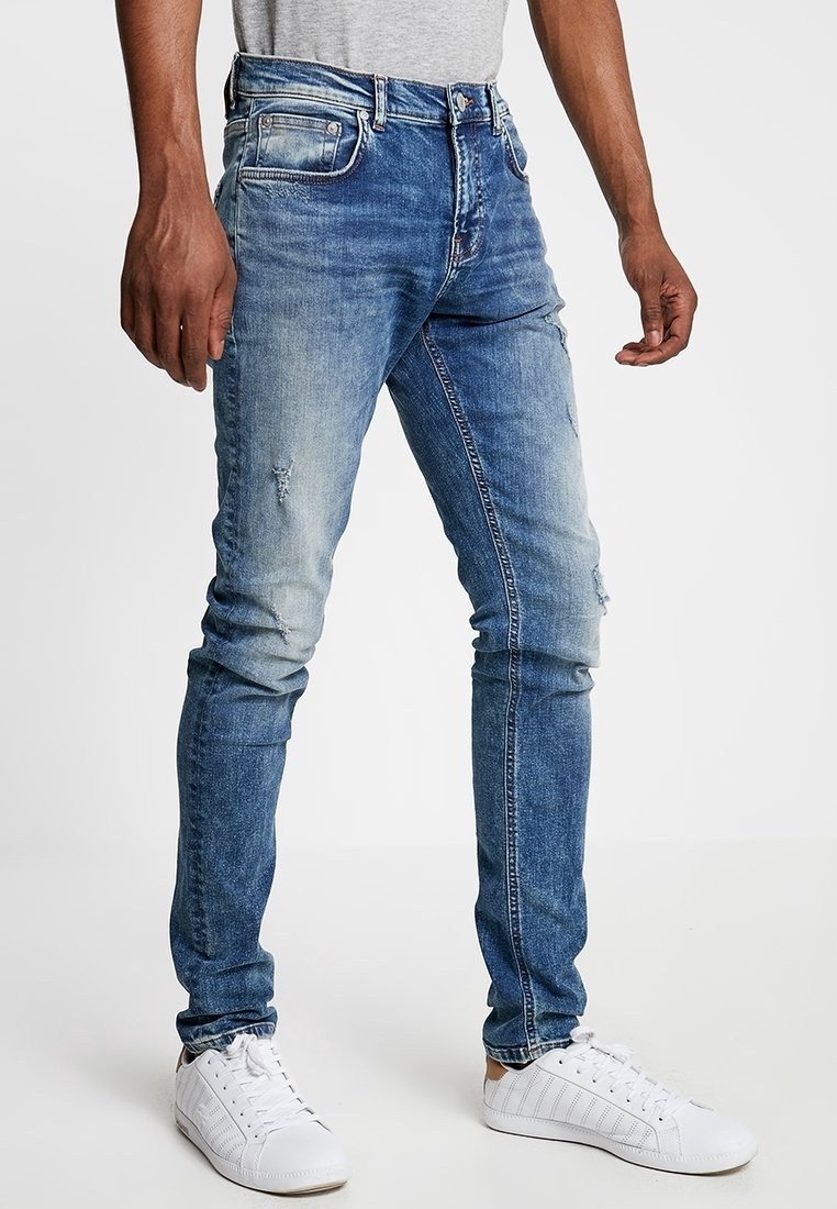 Jean en denim bleu clair avec une coupe slim, présentant des détails usés et un effet délavé. Associé à des baskets blanches et un t-shirt gris.