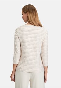 Hellbeige Oberteil mit Dreiviertelärmeln, das ein strukturiertes Wellenmuster aufweist; glattes Material mit Rundhalsausschnitt und lässiger Passform.