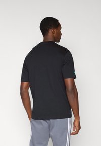 Czarny bawełniany t-shirt z krótkimi rękawami, okrągłym dekoltem i subtelnym zielonym brandingiem na lewym rękawie. Gładka tekstura i standardowy krój.