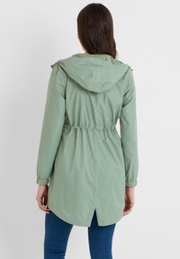 Light green, hooded jacket with elastic waistband and adjustable cuffs. Features a rear vent and a smooth, water-resistant texture.