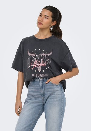 Femme aux longs cheveux foncés en queue de cheval porte un T-shirt noir oversize avec un motif de crâne de taureau et un texte, associé à un jean bleu clair.