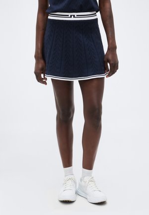 Osoba s tmavou pokožkou, oblečená v námornícky modrej plisovanej sukni s bielym lemovaním, bielych členkových ponožkách a bielych členkových teniskách so šnúrkami, stojaca pred jednofarebným pozadím.