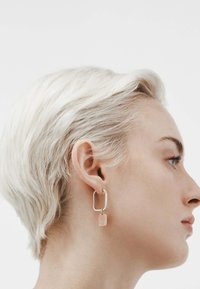 Pendientes de aro cuadrados de plata con una etiqueta de metal rosa que presenta "IVY" en relieve, exhibiendo un diseño geométrico moderno.