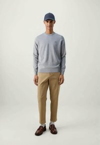 Lys grå genser med ribbestrikkede mansjetter, beige bukser, brune loafers og en mørk blå caps. Modellen står mot en nøytral bakgrunn.