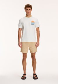 Tricou alb din bumbac cu un grafic colorat de soare și valuri pe piept, asortat cu pantaloni scurți bej și sandale negre.