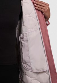 Doublure matelassée d'une veste rose avec une poche latérale ; le tissu extérieur est lisse avec une finition mate. Le tissu présente des motifs de couture.
