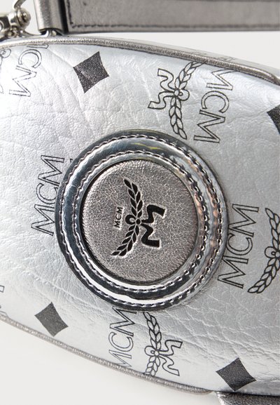 Ασημένια δερμάτινη τσάντα με μοτίβο μαύρου λογότυπου MCM και κυκλικό κεντημένο έμβλημα που απεικονίζει φτερά και τα γράμματα MCM.