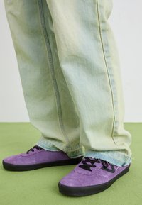 Sapatilhas roxas de camurça com atacadores pretos, combinadas com calças de ganga verde-claro, ligeiramente desgastadas. Close-up no calçado e nas calças.