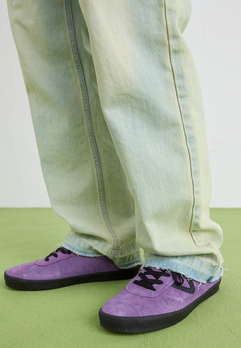 Sapatilhas roxas de camurça com atacadores pretos, combinadas com calças de ganga verde-claro, ligeiramente desgastadas. Close-up no calçado e nas calças.