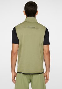 Khaki fleeceväst med hög krage, med en svart logotyp på baksidan, kombinerad med en svart kortärmad skjorta. Slät textur och smal passform.